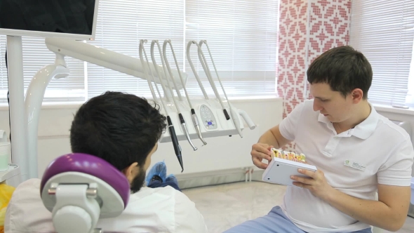 Young Male Dentist Using a Tooth Model Says Patient Is In Chair About Ways Of Treatment.