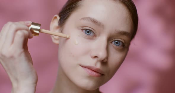 A Young Woman Applying Foundation with a Dropper to Her Cheeks Closeup alt
