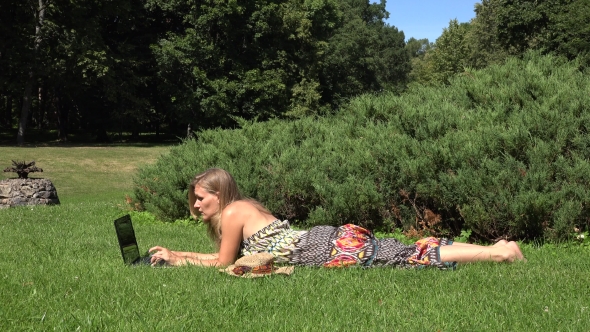 Attractive Female Using Laptop In The Park Lying On The Green Grass.