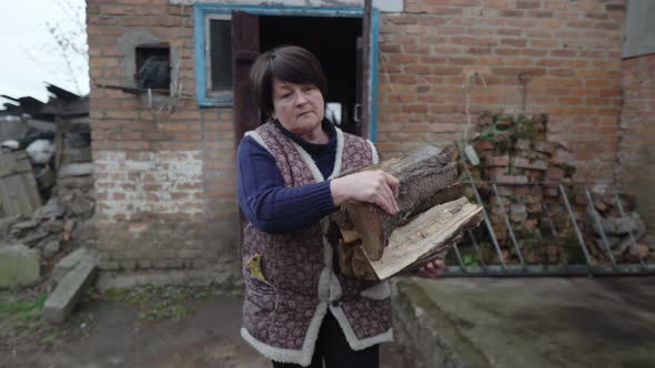 An Elderly Woman with a Handful of Firewood Comes Out of the Barn and Walks Across the Yard