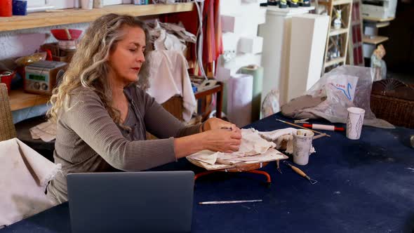 Mature female potter molding a clay while using laptop alt