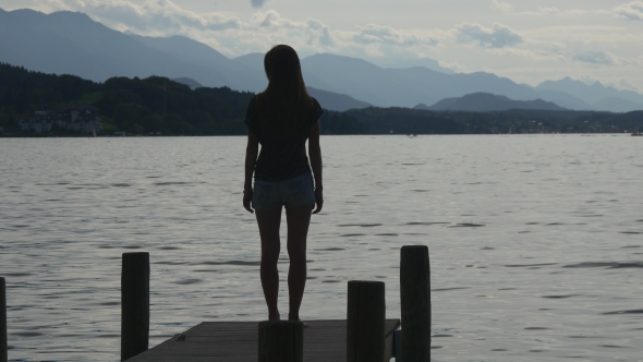 Woman Stands On Pier Of Alpine Lake Alone