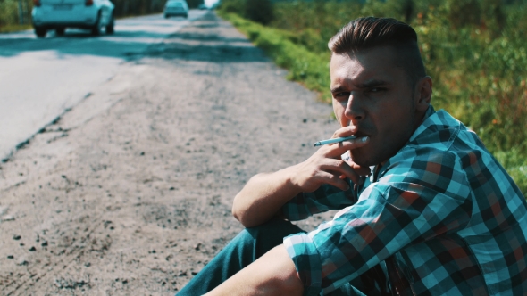 Man Sit At Road In Countryside. Hitchhiking. Waiting. Smoking Cigarette.