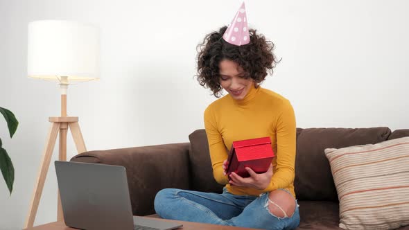 Happy Woman in Party Hat Opens Gift Box and Surprised Sitting in Couch at Home alt
