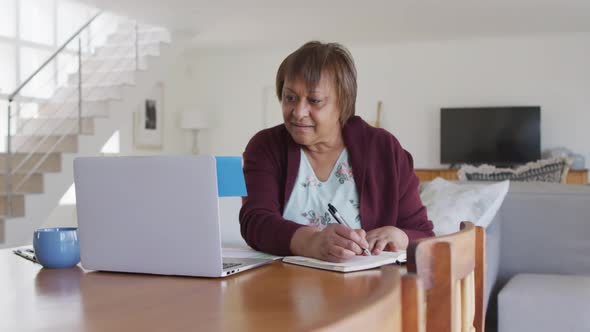 Smiling african american senior woman sitting at dining table, using laptop and writing in notebook alt