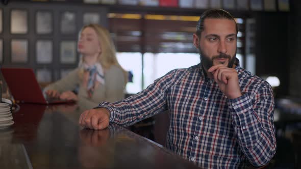Portrait of Handsome Bearded Man Sitting at Bar Counter Waiting for Drink with Blurred Woman Typing alt