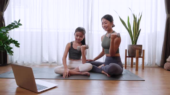 Happy Asian family mother and little daughter exercise by lifting dumbbell follow video from laptop