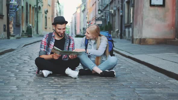 Young Tourists Couple Using Map Sitting on Pavement and Admiring Historical Surroundings alt