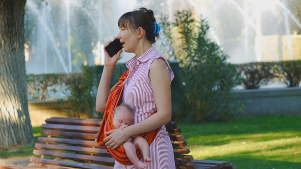 Young Woman With Sleeping Baby in Sling Starts Phone Call alt