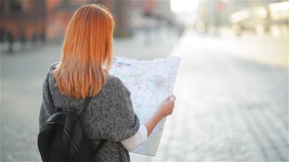 The Young Red-haired Female Tourist Attentively Considers The Map. The Girl Walks Streets alt