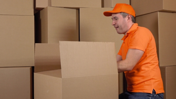 Warehouse Worker In Orange Uniform Packaging Big Carton With Duct Tape Gun. Multiple Boxes alt