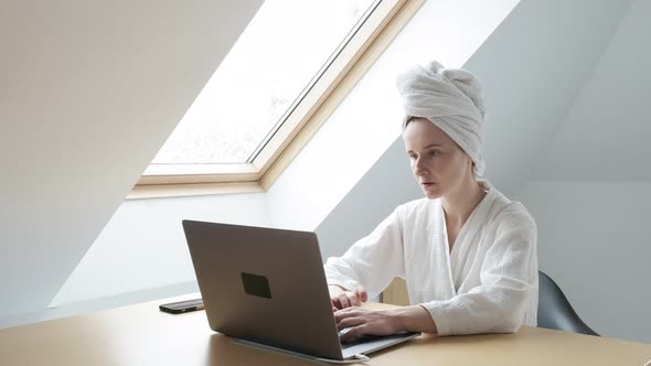 Young Woman in White Bathrobe and Towel on Head Remotely Works at Home on Laptop