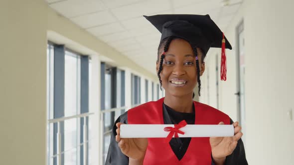 African American Female Graduate in Mantle Stands with a Diploma in Her Hands and Smiles alt