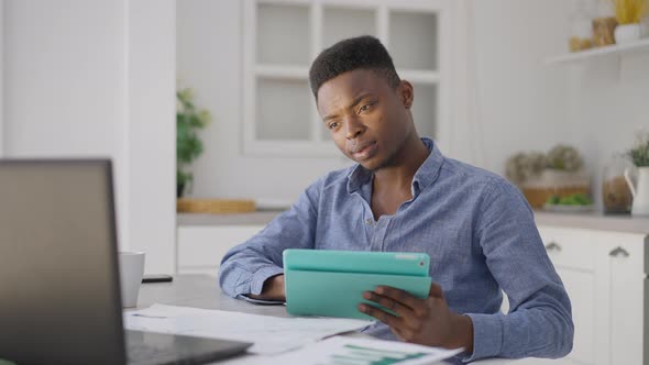 Portrait of Young Handsome Man Scrolling Tablet Screen Checking Laptop Sitting at Kitchen Table alt