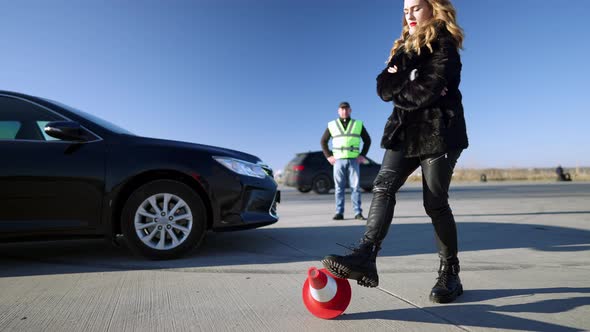 Upset Beautiful Slim Woman Kicking Road Cone with Blurred Auto Instructor Standing at Background alt