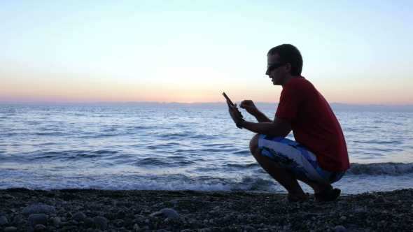 Silhouette Of a Man At Sunset Sitting On The Waterfront With a Tablet Computer
