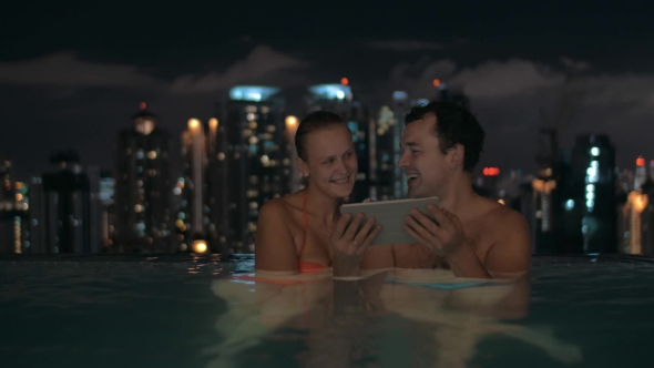 Night View Of Young Beautiful Couple In Swimming Pool Watching In Tablet On The Roof Of Skyscraper alt