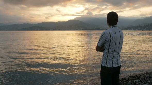 The Man Looks In a Shirt Looking At The Sunrise On The Beach Of The Ocean. The Sun Rises From Behind