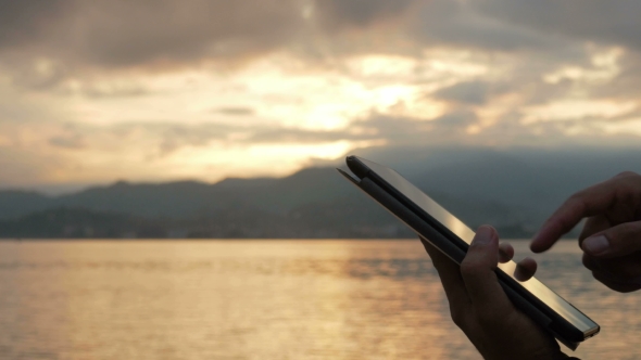 A Man Checks Messages On The Tablet During The Sunrise On The Beach Of The Ocean. Stunning Colors