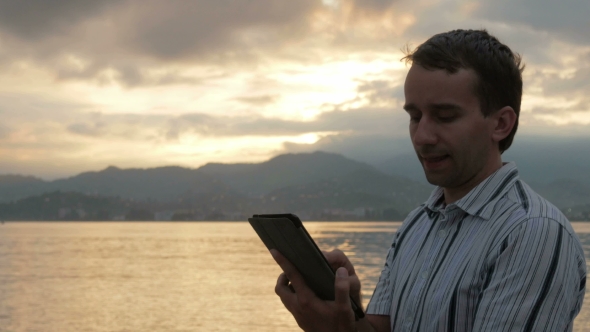 A Man In a Shirt Checks Messages On The Tablet During The Sunrise On The Beach Of The Ocean