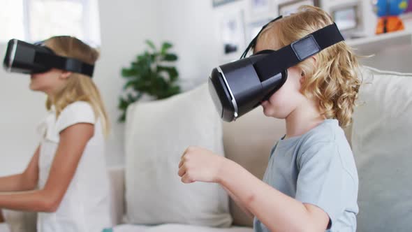 Caucasian brother and sister gesturing while using vr headset sitting on the couch at home alt