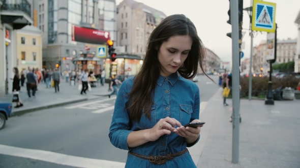 Pretty Woman in a Busy Street. Young Woman in a Blue Dress Standing, Using Her Smartphone alt