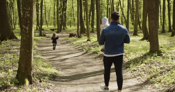 Little Happy Girl Running On A Path In The Woods, Dad With A Child In His Arms Catches Up With Her alt