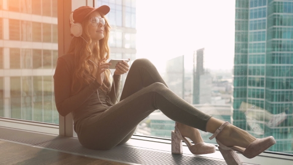 Cute Girl Sitting By Window Listening To Music With Headphones And Sending Sms Using Mobile