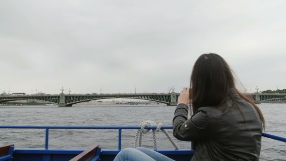 Young Brunette Woman Sightseeing In a River Bus. Taking Photos On a Smartphone Of a Bridge  alt