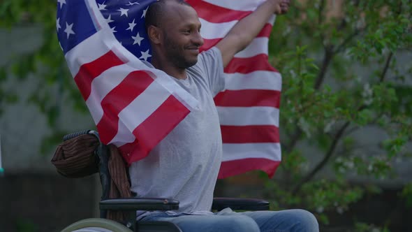 Satisfied Smiling African American Man in Wheelchair Fluttering USA Flag in Hands Looking Away alt