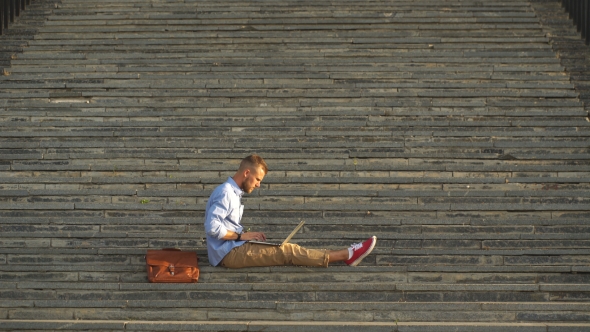 Handsome Student Sitting On Stone Steps And Using a Laptop alt