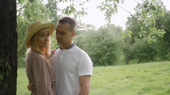 Young Guy with a Woman in a Dress and Hat are Standing By a Tree on a Green Field in Nature in alt