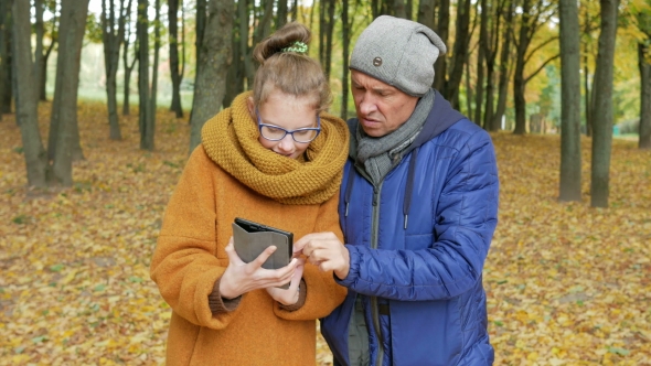 Daughter Teenager Teaches His Father To Work With The Tablet In The Autumn Park. Dad Listens