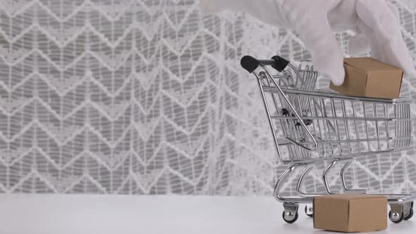 Human Hand in White Cotton Glove Puts Carton Boxes in Mini Shopping Cart alt