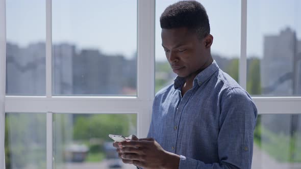 Young Focused Man Typing on Smartphone Screen Standing at Window in Home Office alt