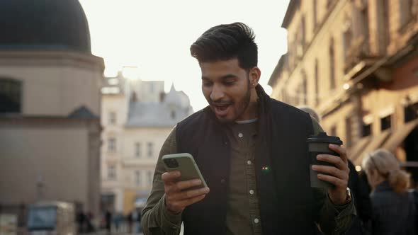 Indian Man with Coffee in Hands Getting Good News on Mobile alt