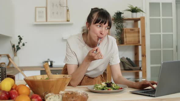 Young Woman Eating Salad and Using Laptop in Kitchen alt