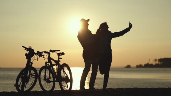 Silhouettes Of Two Cyclists At Sunset. Doing Selfie