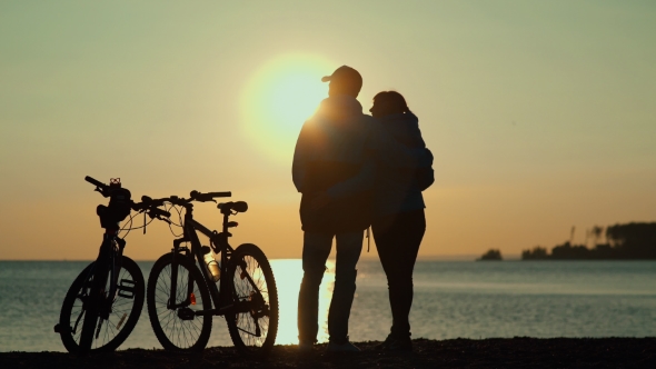The Silhouettes Of Two People With Bicycles On The Beach alt