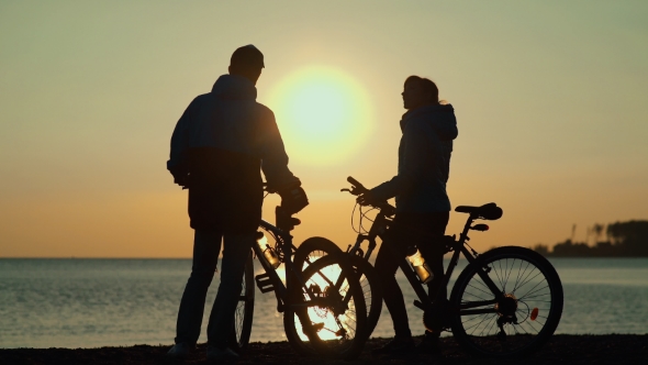 He Silhouettes Of Two People On The Beach With Bikes