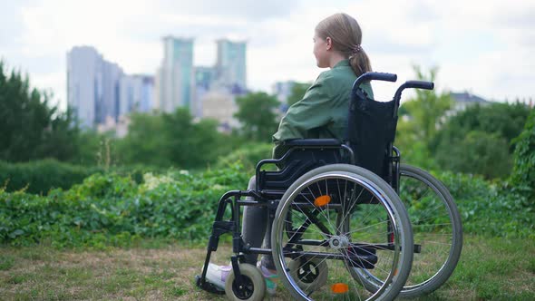 Side View Sad Lonely Teenage Girl in Wheelchair Looking Away Sitting in Summer Park with Urban City alt
