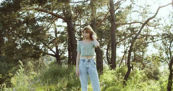 A Cheerful Teenage Girl with Curly Hair Stands in a Clearing Between Green Trees in Summer Forest alt