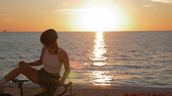 Summertime shot of young woman cyclist in stylish outfit is riding bicycle along beach promenade alt