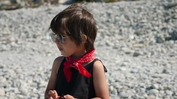 Stylish Child a Boy in a Black Jumpsuit and a Red Scarf Stands on the Beach alt