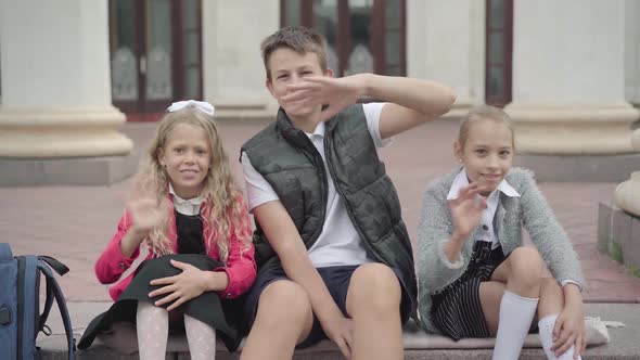 Three Positive Children Waving and Smiling at Camera. Portrait of Cheerful Caucasian Girls and Boy alt