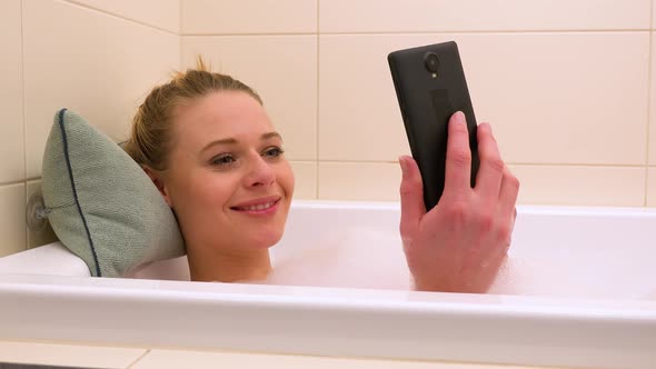A young beautiful woman works on a smartphone in a bathtub with a smile - closeup alt