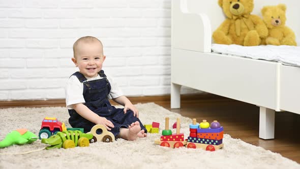 a Baby in a Denim Jumpsuit Sitting Barefoot on the Carpet Smiles and Plays with Toys alt
