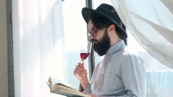 Pleased Bearded Man Drinking Red Wine While Reading a Book at Home alt