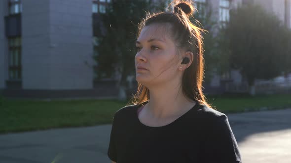 A Woman Is Walking Down the Street with Headphones Towards the Park.