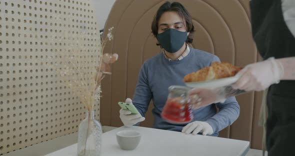 Young Arab Man Wearing Mask Using Smartphone at Cafe Table When Waitress Bringing Food and Drink alt